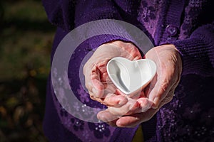 Close-up of a white ceramic heart-shaped on palm senior woman. Valentine`s day. Concept of aged people and healthcare