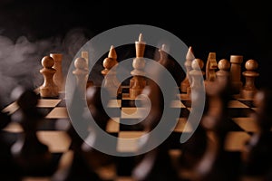 Close up of white chess pieces on board with dissipating steam. Two rows of wooden figures on chessboard on black