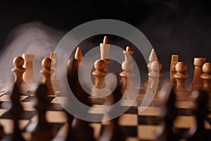 Close up of white chess pieces on board with dissipating steam. Two rows of wooden figures on chessboard on black