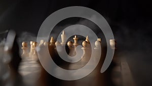 Close up of white chess pieces on board with dissipating steam. Two rows of wooden figures on chessboard on black