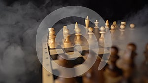 Close up of white chess pieces on board with dissipating steam. Two rows of wooden figures on chessboard on black