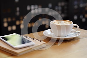 Close up white coffee cup with latte art on wood table with smartphone at coffee shop