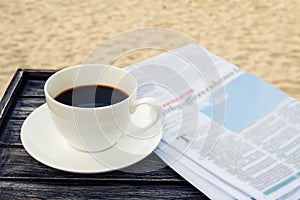 Close up white coffee cup on wood table at sunrise sand beach with newspaper in the morning, warm tone