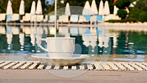 Close-up of a white cup of coffee or hot drink on the background of a swimming pool on a summer sunny day. Good morning