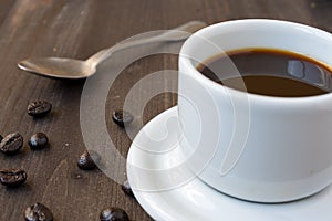 Close-up of white cup with coffee and spoon, on wooden table with coffee beans