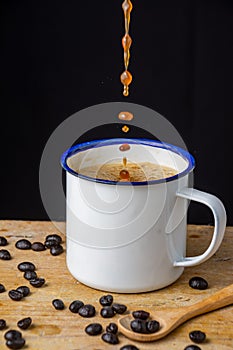 Close-up white cup with falling coffee, coffee beans and spoon on wooden table and black background