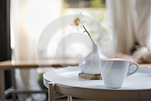 Close up of white cup of hot coffee latte with milk foam on round table with vase of flower in restaurant