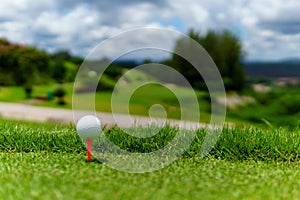 Close up of white golf ball on orange tee on green grass with blue sky and cloud and view of mountain background in sunny day.