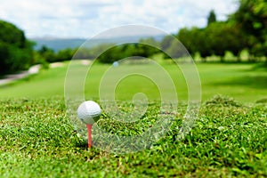 Close up of white golf ball on orange tee on green grass with blue sky and cloud and view of mountain background in sunny day.