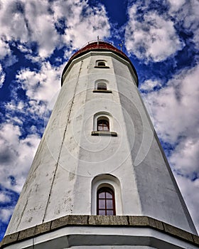 Close up of a white lighthouse with 4 windows on top of each other. Background with blue sky and white clouds. Portrait