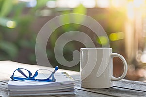 Close up of white mug cup of hot coffee with book and reading glasses on wooden table in garden with copy space