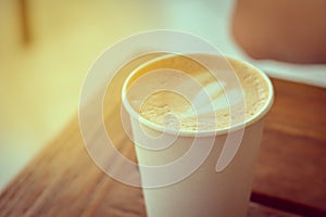 Close up white paper cup of hot coffee cappuccino, latte with heart shape milk foam topping on wooden table with flare light and