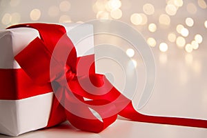 Close up of white paper gift box with a red satin ribbon bow on bright background with golden bokeh. Holidays greeting cards and