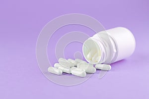 Close up white pill bottle with spilled out capsules on purple background with copy space. Focus on foreground, soft bokeh. Pharma