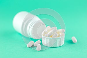 Close up white pill bottle with spilled out pills and capsules in cap on aquamarine background with copy space. Focus on foregroun