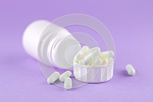 Close up white pill bottle with spilled out pills and capsules in cap on purple background with copy space. Focus on foreground, s