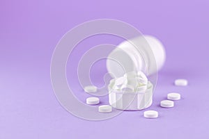 Close up white pill bottle with spilled out pills and capsules in cap on purple background with copy space. Focus on foreground, s