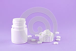 Close up white pill bottle with spilled out pills and capsules in cap on purple background with copy space. Focus on foreground, s