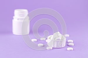 Close up white pill bottle with spilled out pills and capsules in cap on purple background with copy space. Focus on foreground, s