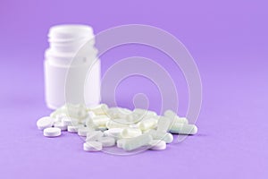 Close up white pill bottle with spilled out pills and capsules on purple background with copy space. Focus on foreground, soft bok