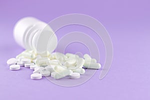 Close up white pill bottle with spilled out pills and capsules on purple background with copy space. Focus on foreground, soft bok