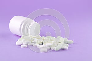 Close up white pill bottle with spilled out pills and capsules on purple background with copy space. Focus on foreground, soft bok