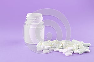 Close up white pill bottle with spilled out pills and capsules on purple background with copy space. Focus on foreground, soft bok