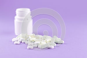 Close up white pill bottle with spilled out pills and capsules on purple background with copy space. Focus on foreground, soft bok