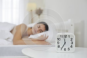 Alarm clock set for 7 AM on bedside table, with happy woman sleeping on bed in background photo