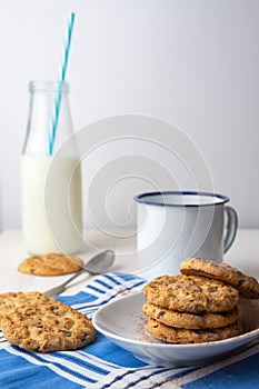 Close-up of white plate with chocolate chip cookies, cup, spoon and bottle of milk with blue straw, on blue napkin and white backg