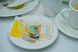 Close-up of white plate for money, Swiss francs banknotes and coins, Restaurant bill, cup of coffee, delicate pink flowers, dishes