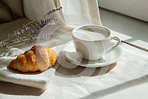 Close-up of a white saucer with cup of cappuccino and brioche on a white linen tablecloth. Classic Italian breakfast set