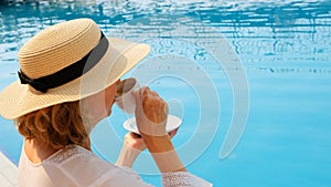 Close up of a woman aged 50-55 in a straw hat drinking from a cup of coffee next to a blue swimming pool, luxurious good