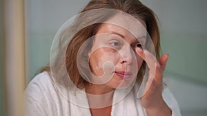 Close-up. A woman applies cream to her cheek and nose with her finger, while sitting in a white bathrobe in front of a