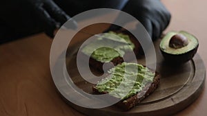 Close-up of Woman in black glowes puts guacamole or avocado spread on top of rye bread toast on wooden board at home