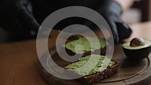 Close-up of Woman in black glowes puts guacamole or avocado spread on top of rye bread toast on wooden board at home