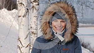 Close-up woman in blue down jacket with fur hood frown from the sun and smiles,in winter Park