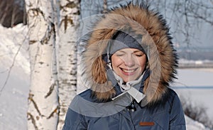 Close-up woman in blue down jacket with fur hood frown from the sun and smiles,in winter Park.