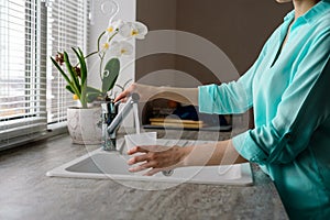 Close-up of a woman collects water in a plastic glass from the tap in the kitchen sink in front of the window