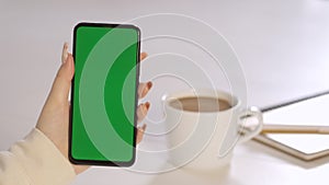 Close-up of a woman drinking coffee and using a phone with a green screen sitting at a wooden table.business woman using