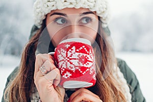 Close up of woman drinking hot tea holding cup dressed in red knitted Christmas case in snowy winter park.