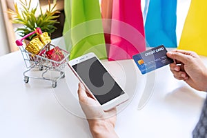 Close up of woman hand holding credit card while using smartphone with miniature gift boxes in trolley and colorful bags on white