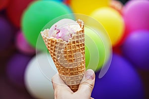 Close up of woman hand holding fresh waffle cone with strawberry ice cream on the background of colorful balloons