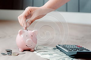 Close-Up Woman Hand is Putting a Coin into Piggy Bank and Calculator and Money Cash on The Bedroom., Female Hand is Inserting Coin