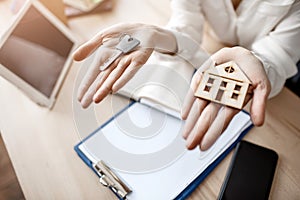 Close up woman hands holding key and wooden house. Documents tablet and phone on table.