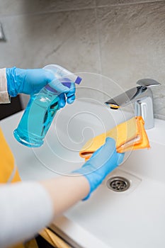 Close up of woman hands in protective gloves cleaning bathroom faucet tap and basin with cloth and sanitizer spray. Hygiene