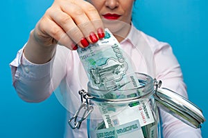 Close-up woman holds a jar in her hands with paper money stashes on a blue background.