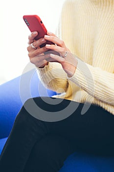 Close-up of woman in pale yellow jumper sitting on blue sofa and holding in hands modern smartphone in red case.