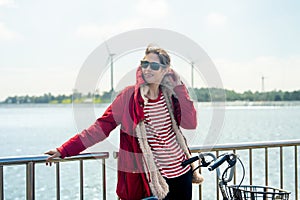 Close up Woman with red coat and sun glasses lean to fence and touch coat hood also hold hand of bicycle stand in front of wind