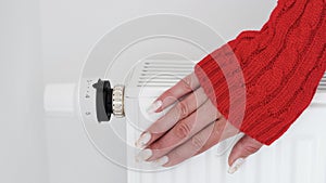Close-up of a woman in red sweater warms her hands over the battery on background of white wall.Macro photo of woman`s hands,copy